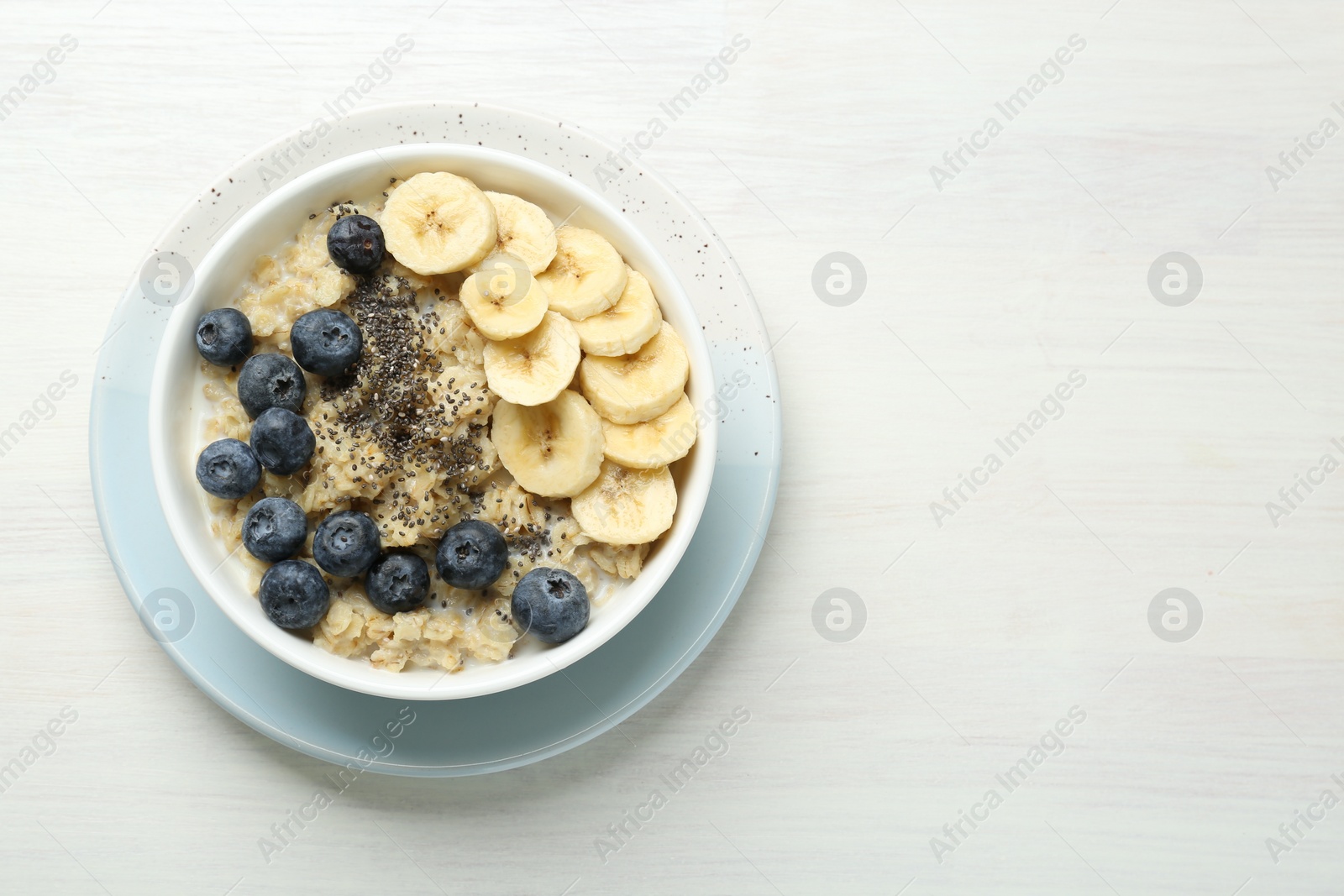 Photo of Tasty oatmeal with banana, blueberries and chia seeds served in bowl on white wooden table, top view. Space for text