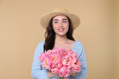 Photo of Beautiful young woman in straw hat with bouquet of pink peonies against beige background