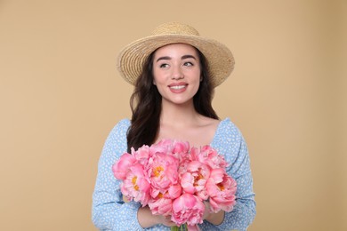 Beautiful young woman in straw hat with bouquet of pink peonies against beige background