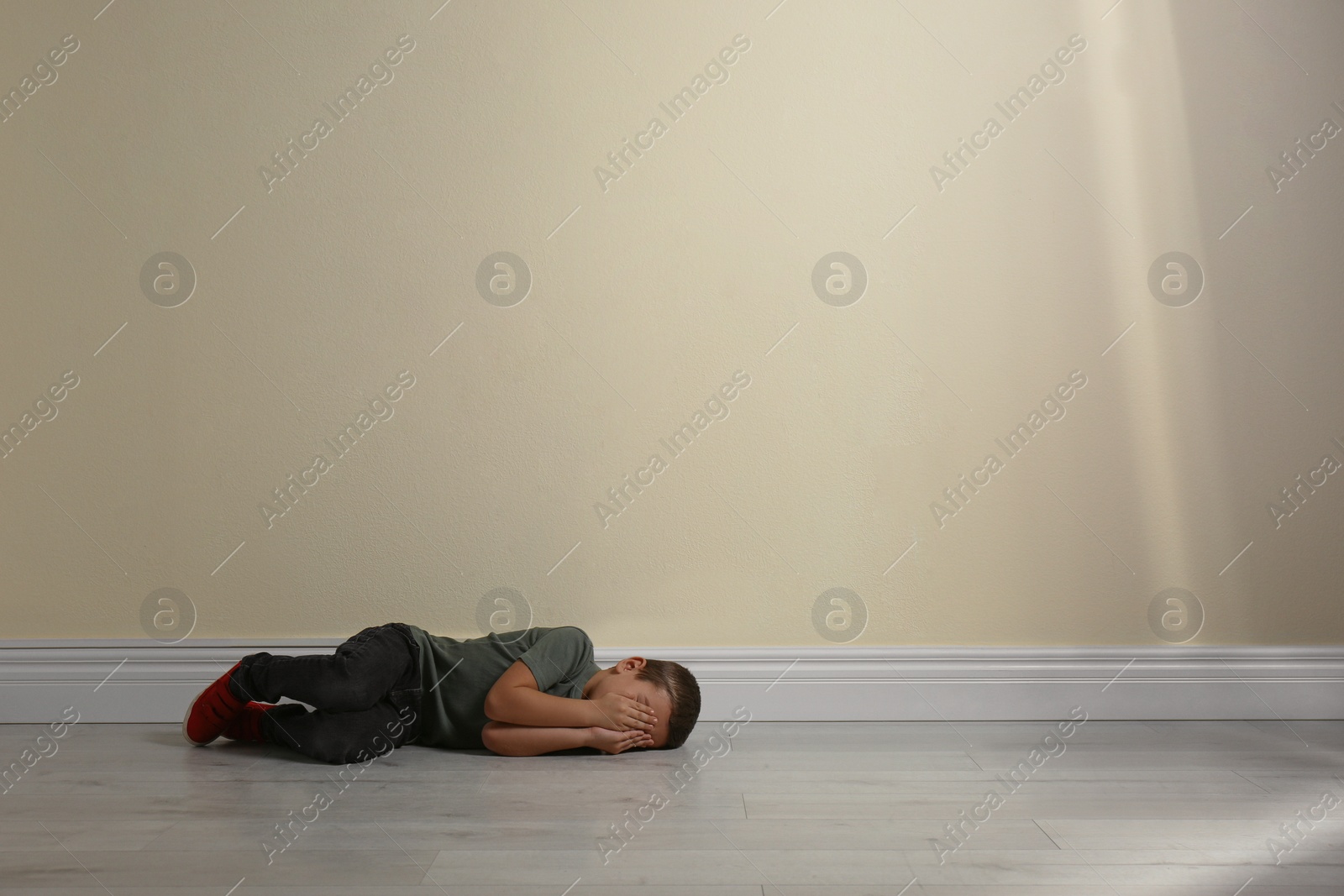 Photo of Little boy closing face with hands lying on floor near yellow wall, space for text. Child in danger