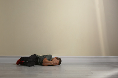 Photo of Little boy closing face with hands lying on floor near yellow wall, space for text. Child in danger