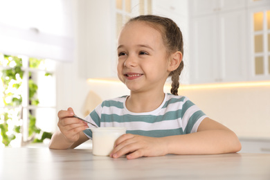 Photo of Cute little girl with tasty yogurt at table in kitchen