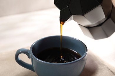 Pouring aromatic coffee from moka pot into cup at white table, closeup