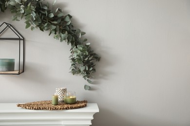 Photo of Beautiful garland made of eucalyptus branches and aromatic candle hanging above mantelpiece indoors