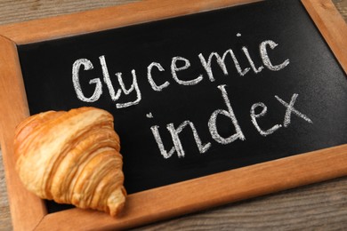Photo of Small chalkboard with words Glycemic Index and croissant on wooden table, closeup