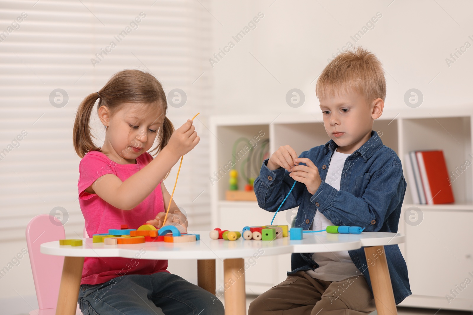Photo of Little children playing with wooden pieces and string for threading activity at white table indoors. Developmental toys