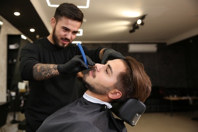 Photo of Professional hairdresser shaving client with straight razor in barbershop