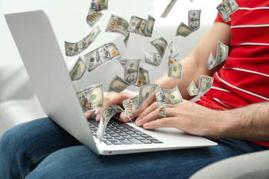 Man with modern laptop and flying dollar banknotes at home, closeup. People make money online