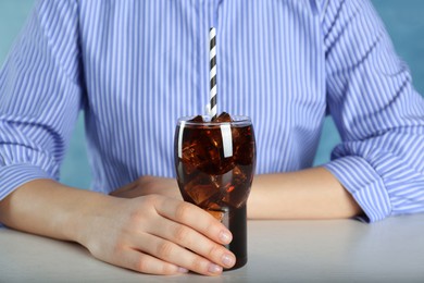 Photo of Woman holding glass of cola with ice and straw at white table against light blue background, closeup. Refreshing soda water