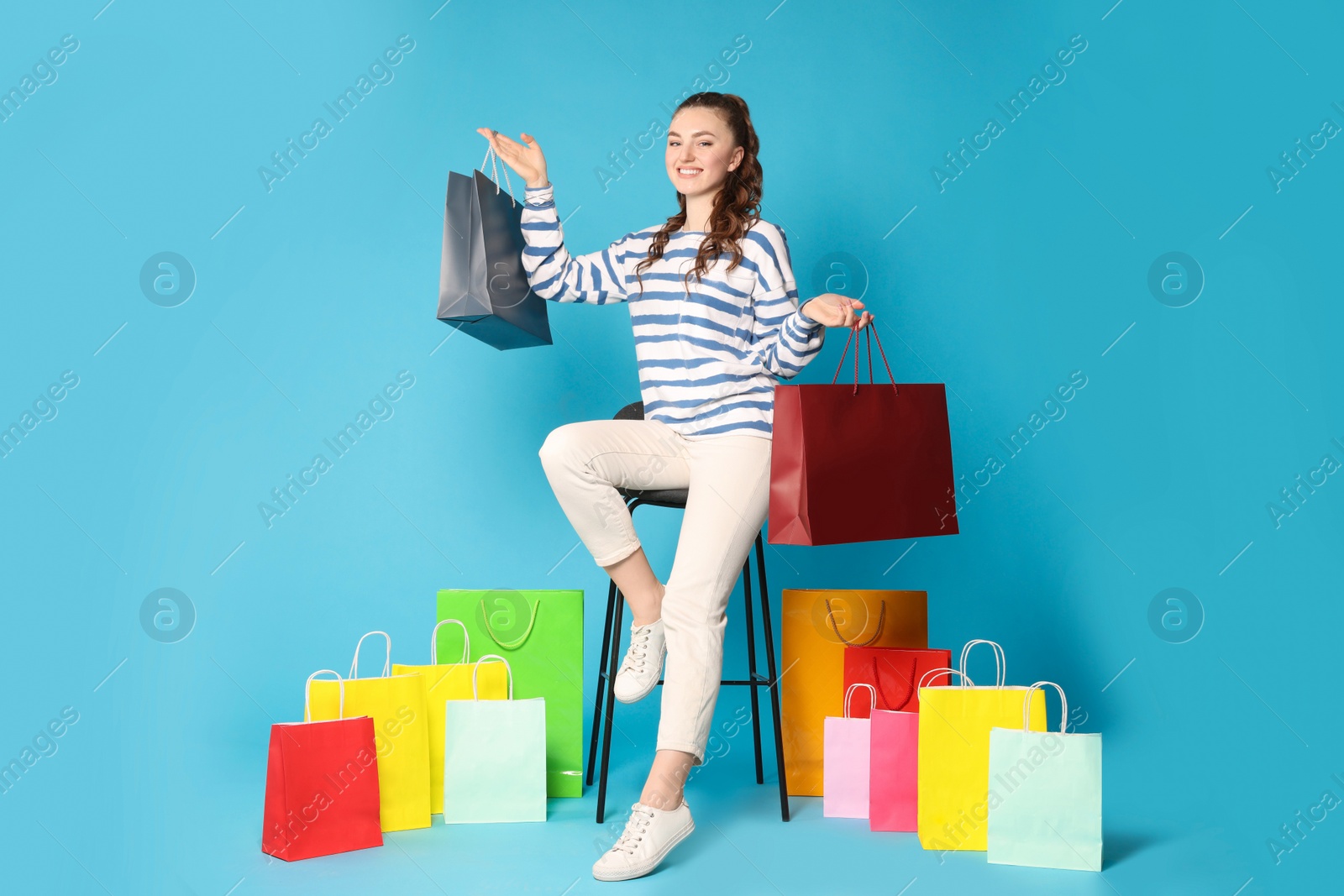 Photo of Happy woman holding colorful shopping bags on stool against light blue background