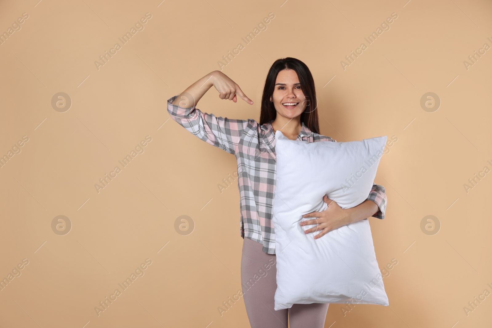 Photo of Happy young woman pointing at soft pillow on beige background, space for text