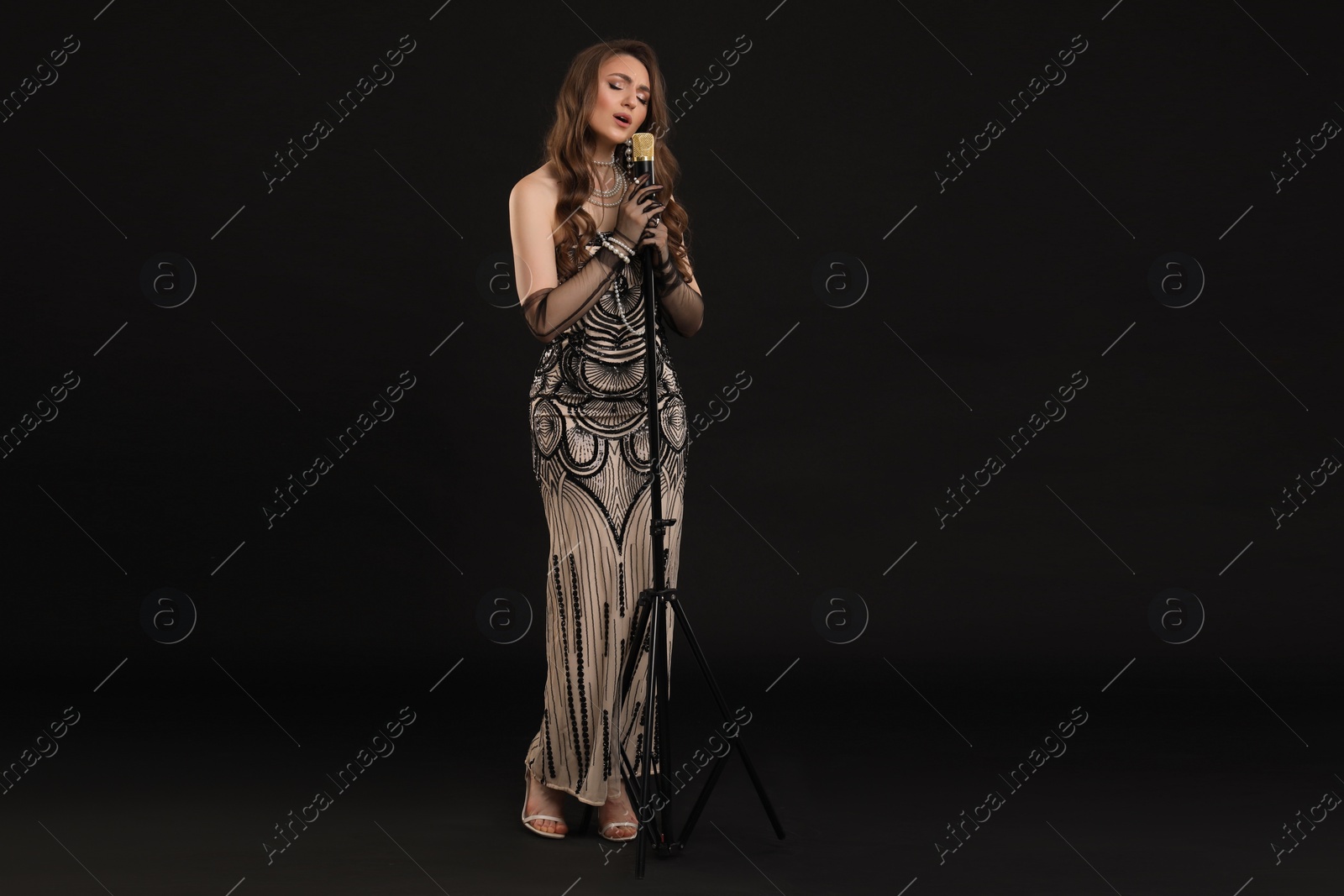 Photo of Beautiful young woman in stylish dress with microphone singing on black background, space for text