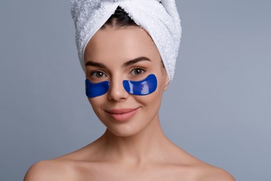 Beautiful young woman with under eye patches and hair wrapped in towel on grey background