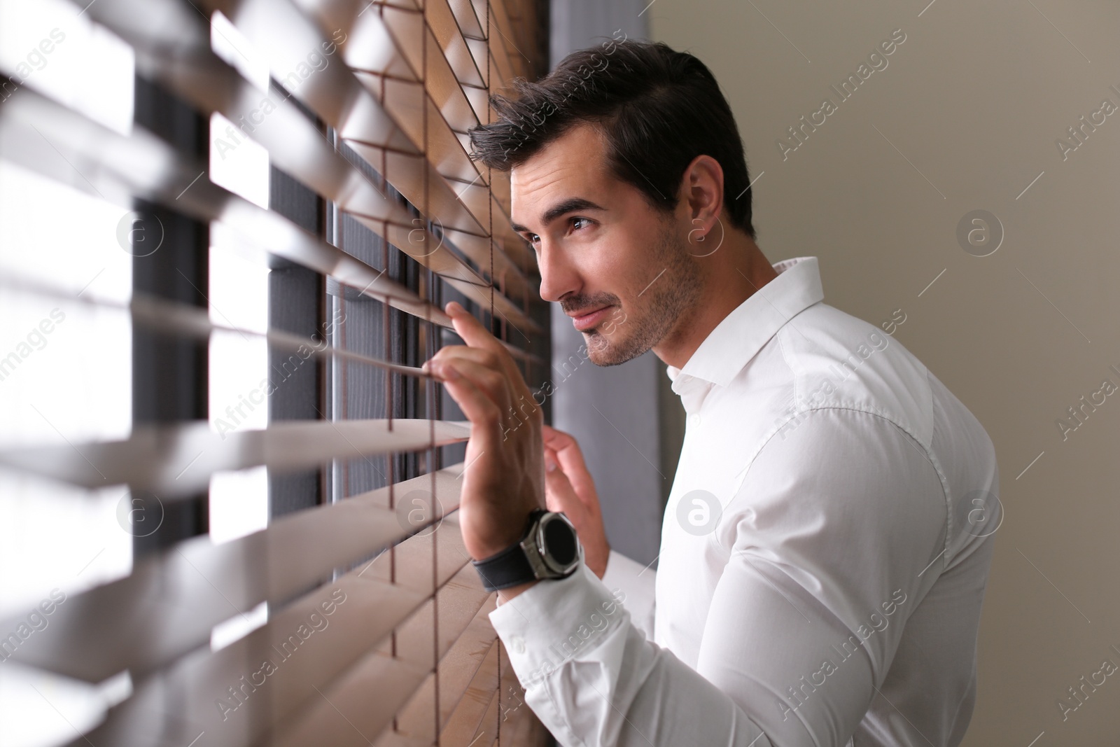 Photo of Handsome young man looking through window blinds indoors