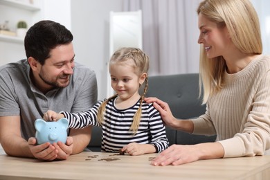 Family budget. Little girl putting coin into piggy bank while her parents watching indoors