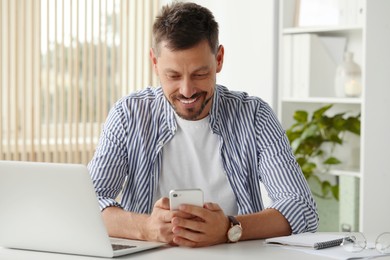 Happy man using smartphone while working with laptop in office