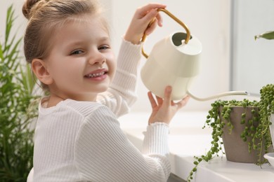 Cute little girl watering beautiful green plant on windowsill at home. House decor