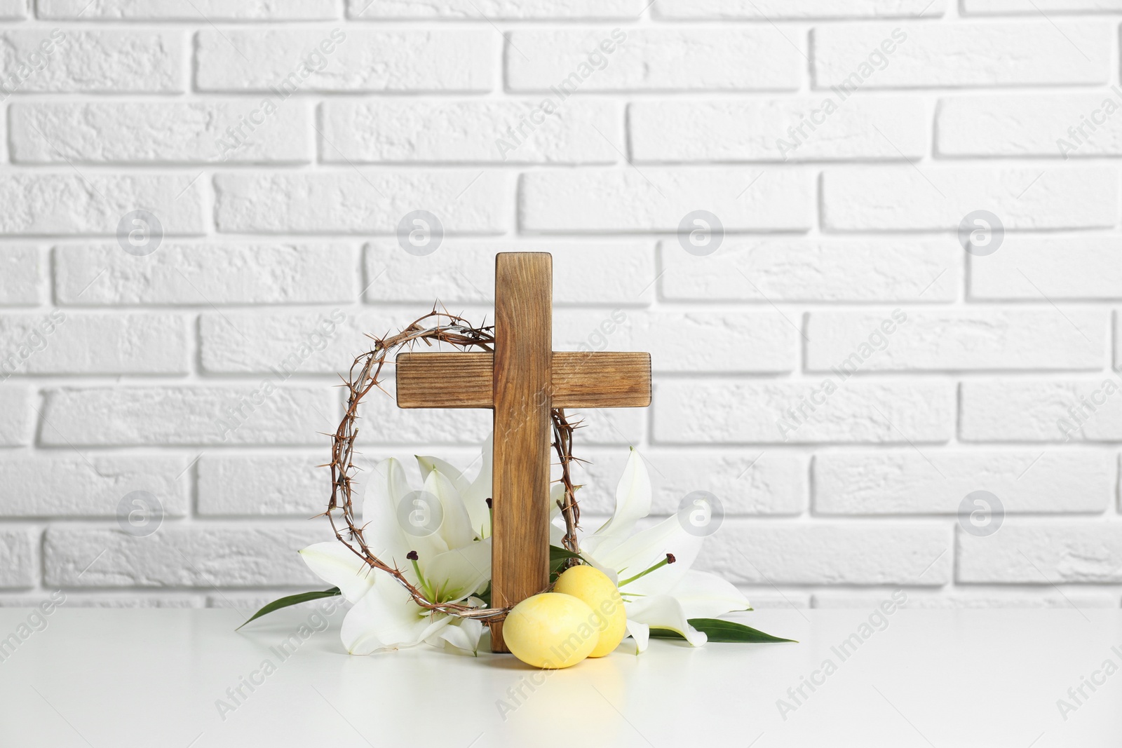 Photo of Wooden cross, crown of thorns, Easter eggs and blossom lilies on table against brick wall