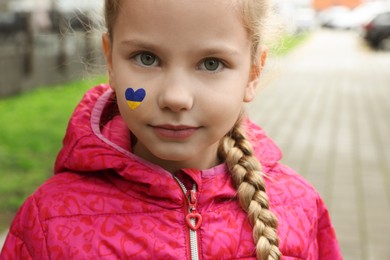 Photo of Little girl with drawing of Ukrainian flag on face in heart shape outdoors