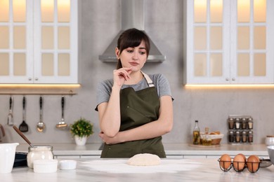 Pensive young housewife with raw dough at white marble table in kitchen. Cooking process