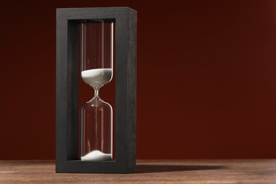 Photo of Hourglass with flowing sand on wooden table against brown background, space for text