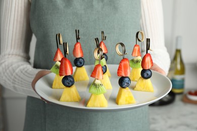 Photo of Woman holding plate of tasty canapes with pineapple, kiwi and berries indoors, closeup