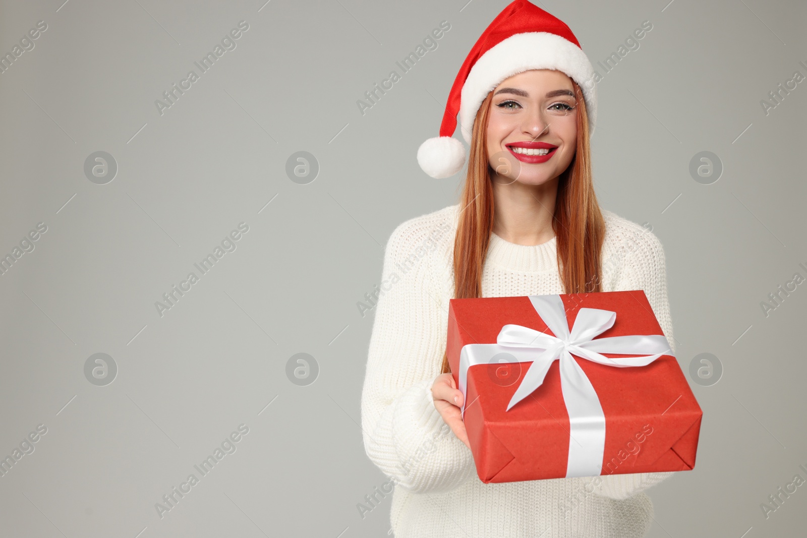 Photo of Young woman in Santa hat with Christmas gift on light grey background, space for text