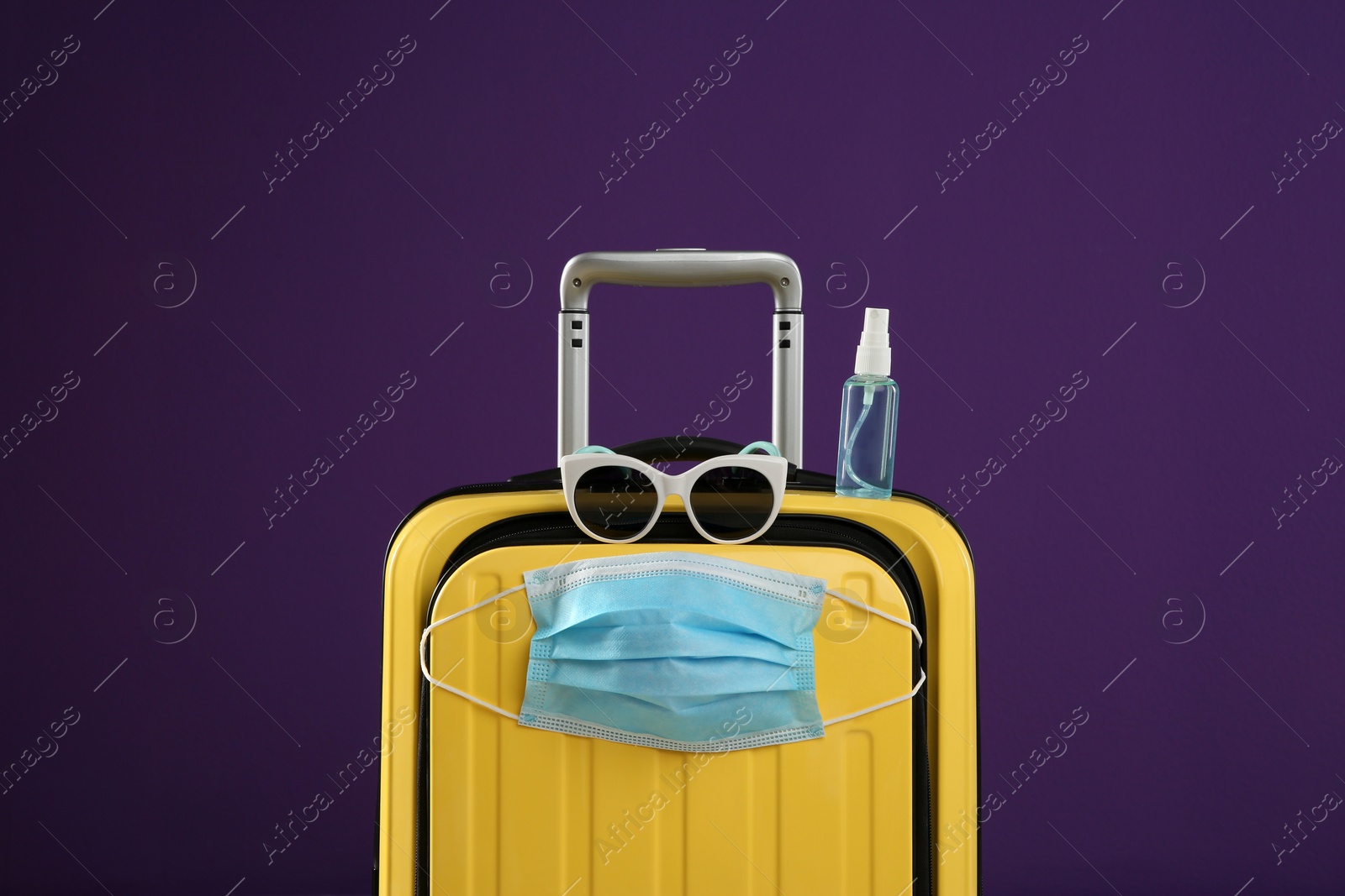 Photo of Stylish yellow suitcase with protective mask, sunglasses and antiseptic spray on purple background. Travelling during coronavirus pandemic