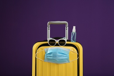 Photo of Stylish yellow suitcase with protective mask, sunglasses and antiseptic spray on purple background. Travelling during coronavirus pandemic