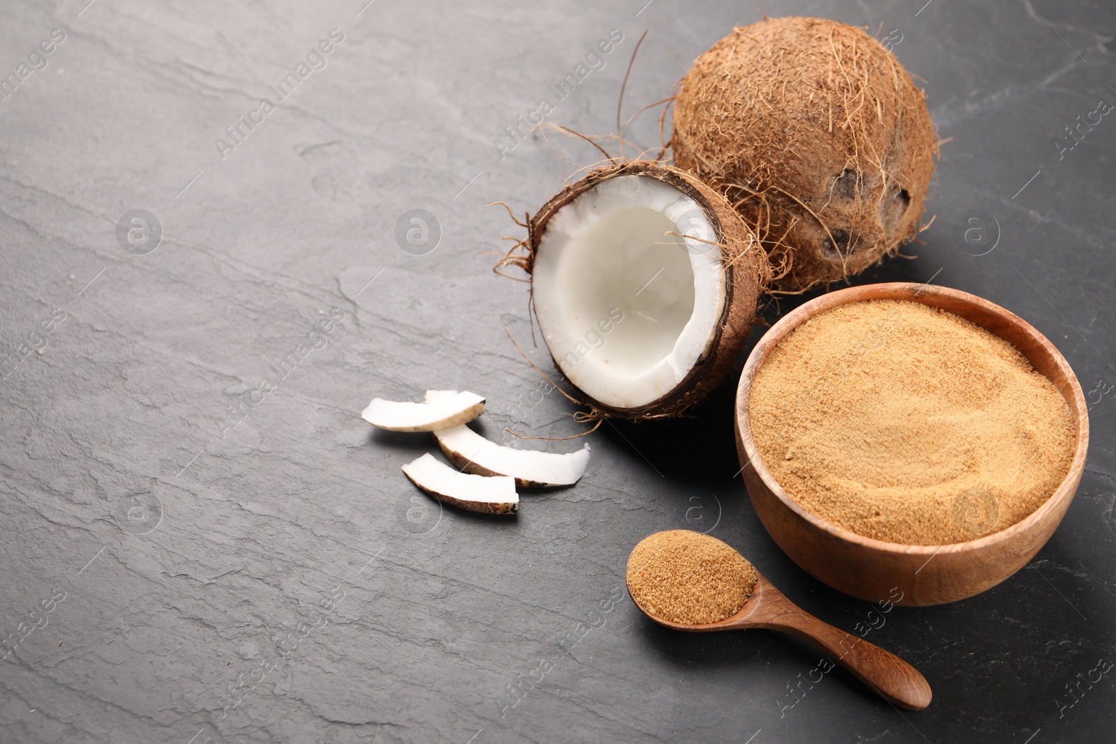 Photo of Spoon with coconut sugar, bowl and fruits on dark textured table, closeup. Space for text