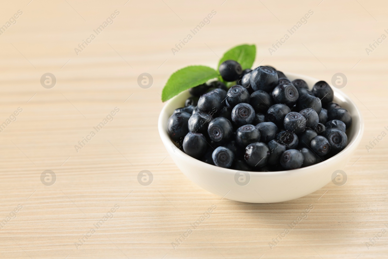 Photo of Tasty fresh bilberries with leaves in bowl on light table. Space for text