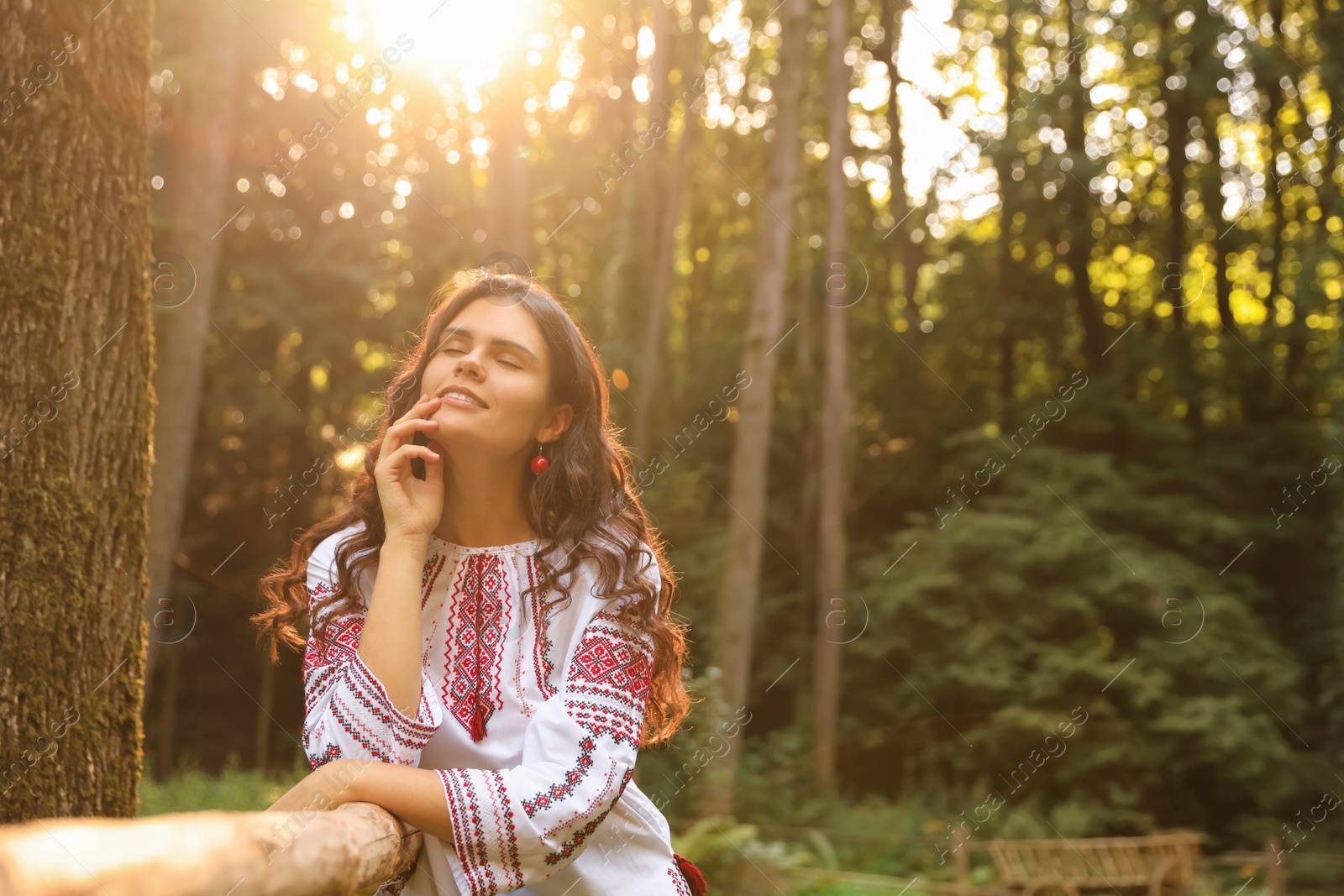 Photo of Beautiful woman wearing embroidered shirt near wooden railing in countryside, space for text. Ukrainian national clothes