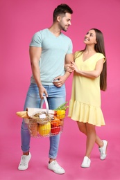 Young couple with shopping basket full of products on pink background