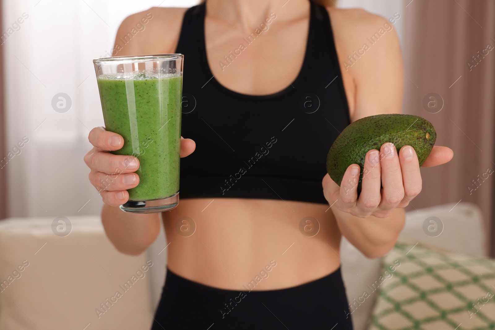 Photo of Young woman in sportswear with glass of fresh smoothie and avocado at home, closeup