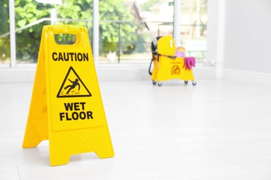 Photo of Safety sign with phrase "CAUTION WET FLOOR" and mop bucket on floor, indoors. Cleaning tools