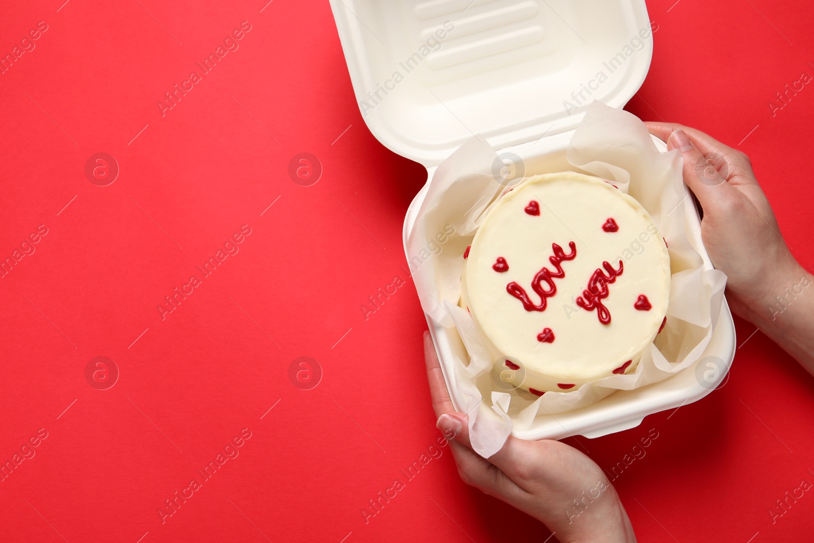 Photo of Woman holding takeaway box with bento cake at red table, top view. St. Valentine's day surprise