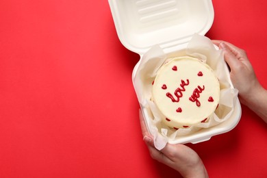 Photo of Woman holding takeaway box with bento cake at red table, top view. St. Valentine's day surprise