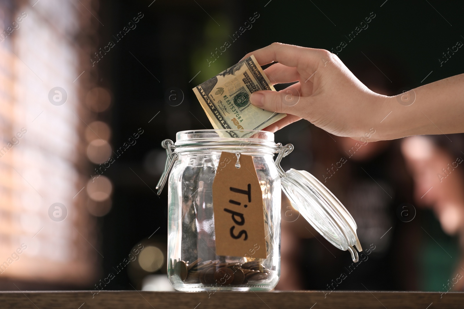 Photo of Woman putting tips into glass jar on wooden table indoors, closeup