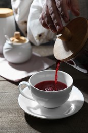 Woman pouring freshly brewed hibiscus tea from teapot into cup at wooden table, closeup