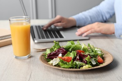 Photo of Office employee working with laptop at white wooden table, focus on fresh vegetable salad with glass of juice. Business lunch