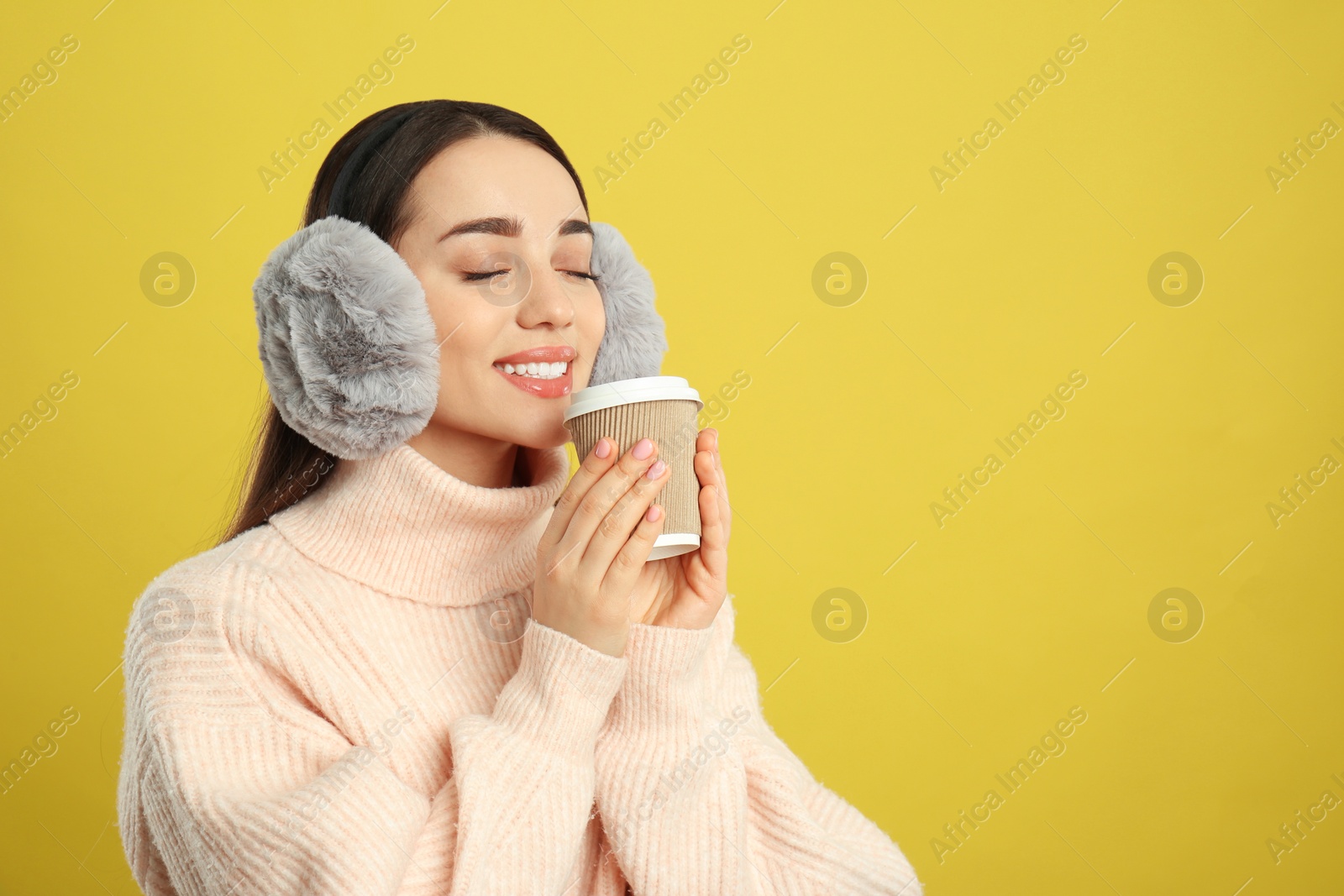 Photo of Beautiful young woman in earmuffs with cup of drink on yellow background. Space for text