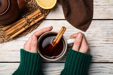 Woman holding cup of delicious hot tea with cinnamon at white wooden table, top view