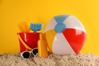 Photo of Colorful inflatable ball, plastic beach toys, sunscreen and sunglasses on sand against yellow background
