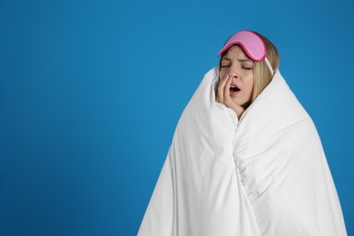 Young woman in sleeping mask wrapped with soft blanket yawning on blue background. Space for text