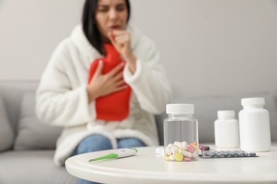 Photo of Ill woman with hot water bottle coughing at home, focus on pills