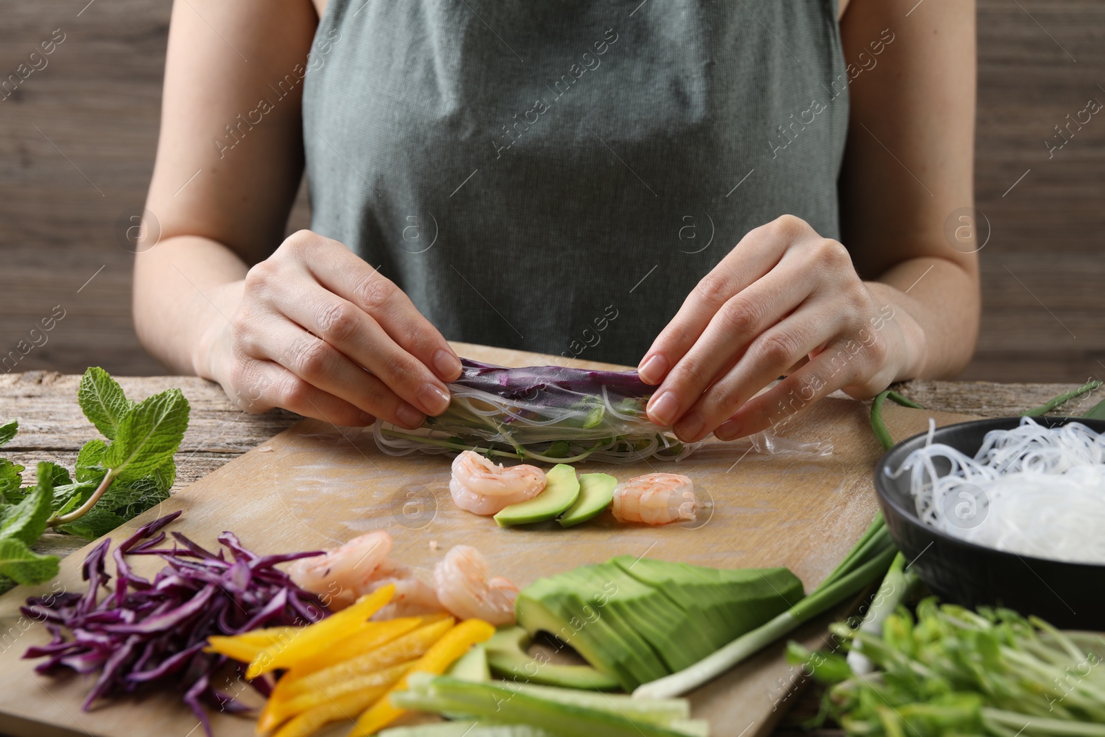 Photo of Woman wrapping spring roll at wooden table with products, closeup