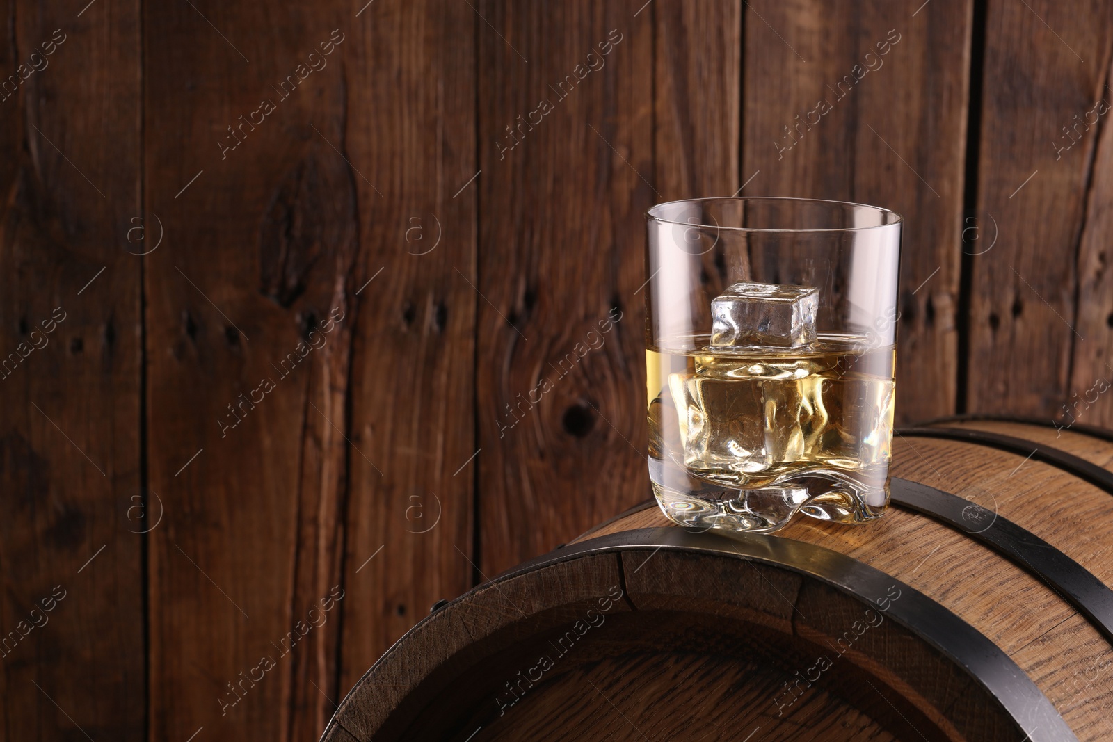 Photo of Whiskey with ice cubes in glass on barrel against wooden background, space for text