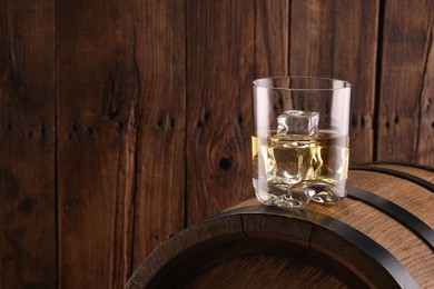 Whiskey with ice cubes in glass on barrel against wooden background, space for text