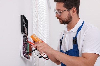 Photo of Technician using digital multimeter while installing home security alarm system on white wall indoors