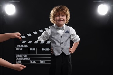 Photo of Smiling cute boy performing while second assistant camera holding clapperboard on stage. Little actor